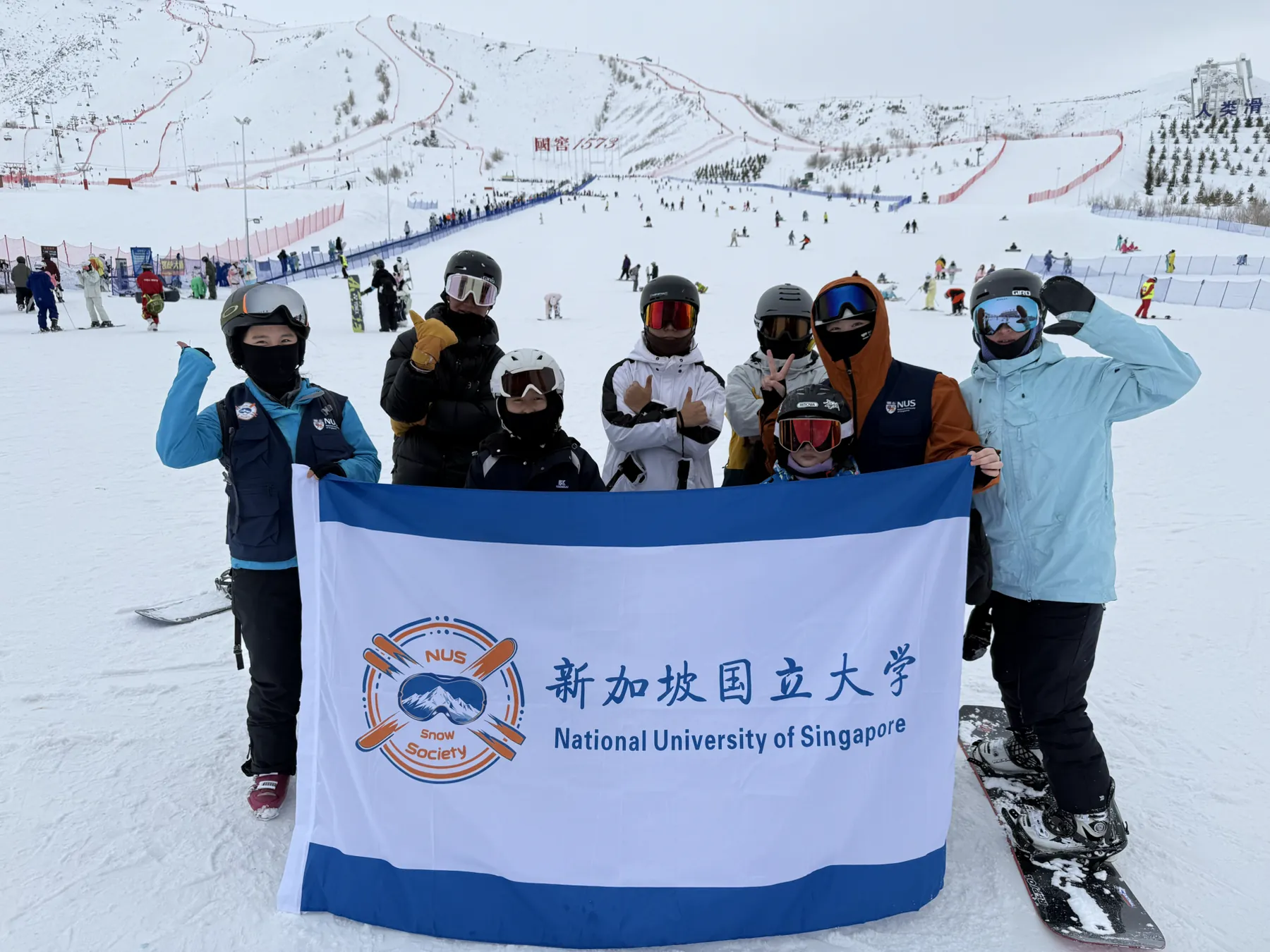 NUS Snow Society members in ski and snowboard gear holding an NUS banner on a snowy slope in Xinjiang
